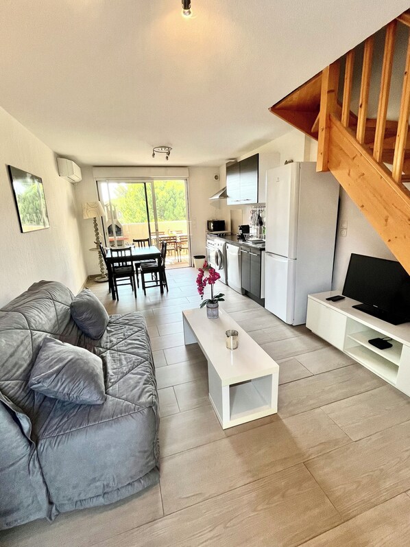 Ein modernes Wohnzimmer mit einem grauen Sofa, einem weißen Couchtisch und einem Fernsehschrank. Es gibt eine Küchenzeile mit einer Waschmaschine sowie einen Essbereich mit Tisch und Stühlen.