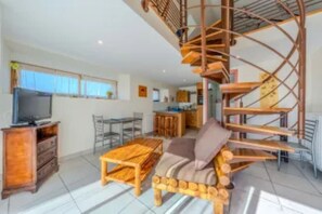 A modern interior with a wooden sofa, a television, a dining area, and a spiral staircase. - Gîte Azalée Romantique 2 persons (Plouarzel)