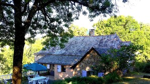 Une maison en pierre avec un toit en pente, entourée d’arbres et d’une zone herbeuse.