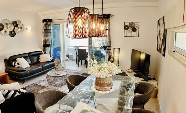 A modern living room with a black leather sofa, a glass dining table, and a large TV.