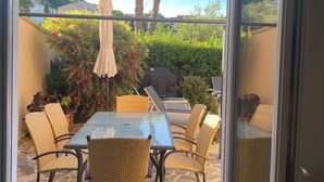 Een terras met een glazen deur, rieten stoelen, een glazen tafel en een barbecue.