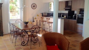 Een eethoek met een tafel en stoelen, een keuken met moderne apparatuur, en een rieten stoel met een rode kussens.