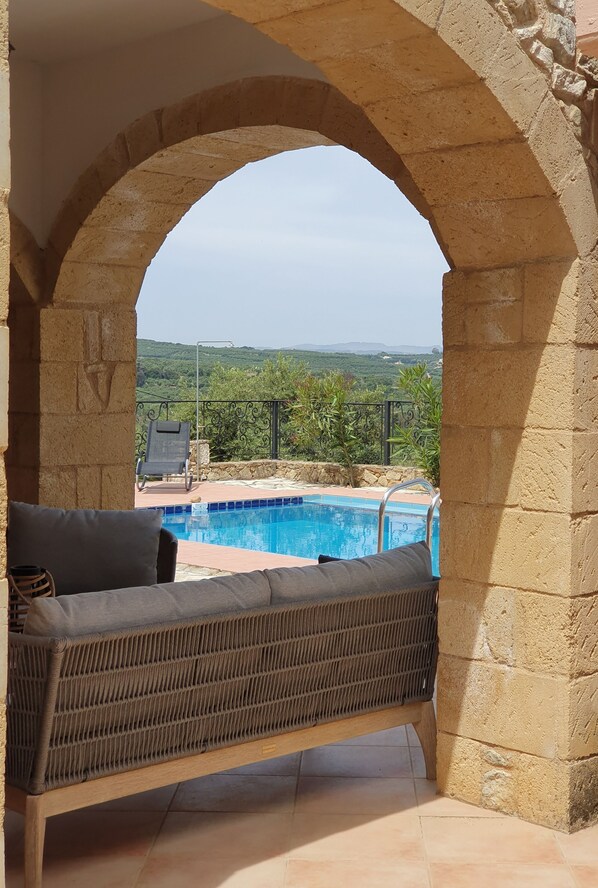 A stone archway leading to a pool area with a wicker sofa and a view of greenery. - House located in the center of an exceptional region to discover ! (Platanias Pirgos Spilorenou Crète)