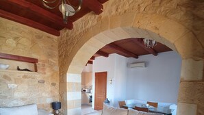 A cozy living room with a stone wall, wooden ceiling beams, a sofa, a coffee table, and a kitchen in the background. - House located in the center of an exceptional region to discover ! (Platanias Pirgos Spilorenou Crète)