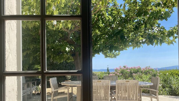 A view through a window of a patio area with a table and chairs, surrounded by greenery and a view of the landscape.