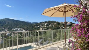 A poolside area with a view of a hillside town, a metal railing, and a beige umbrella.
