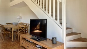 A living room with a TV on a wooden stand, a dining table with chairs, and a staircase with white railings.