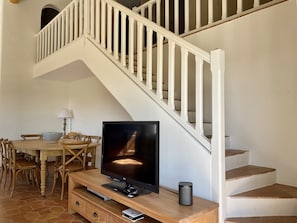 Una sala de estar con un televisor sobre un mueble de madera, una mesa de comedor con sillas y una escalera con barandillas blancas.