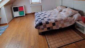 A bedroom with a sloped ceiling, wooden flooring, a bed with a checkered duvet, a bedside table, a nightstand, a window, and a storage unit with colored compartments.