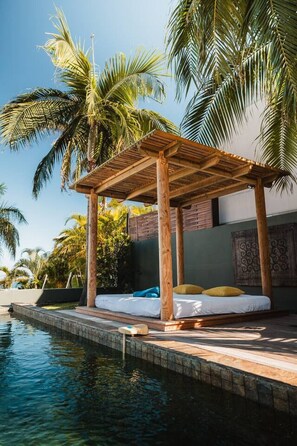 A wooden gazebo with a bed, a pool, and palm trees.