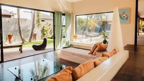 A modern living room with a glass coffee table, a sofa with cushions, a black chair, and a view of a garden through large windows.