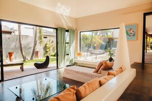 Una sala de estar moderna con una mesa de centro de vidrio, un sofá con cojines, una silla negra y vistas a un jardín a través de ventanas grandes.