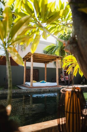 Un quiosco de madera sobre una piscina, con una cama y almohadas, rodeado de follaje.