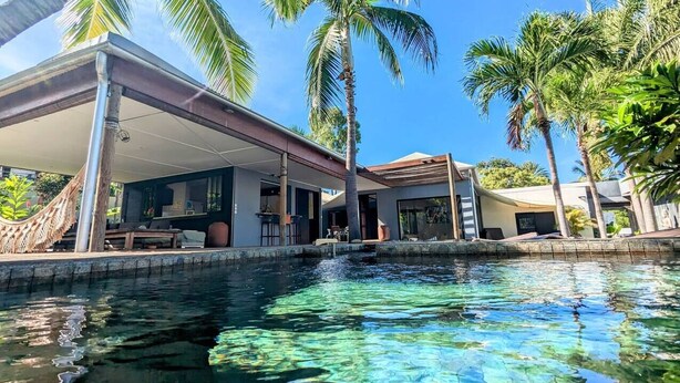 A pool with clear water, a covered outdoor seating area, and palm trees.