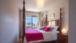 Une chambre à coucher avec un lit à baldaquin, une table de chevet avec une lampe, une fenêtre avec des rideaux et une vue sur les bâtiments environnants.