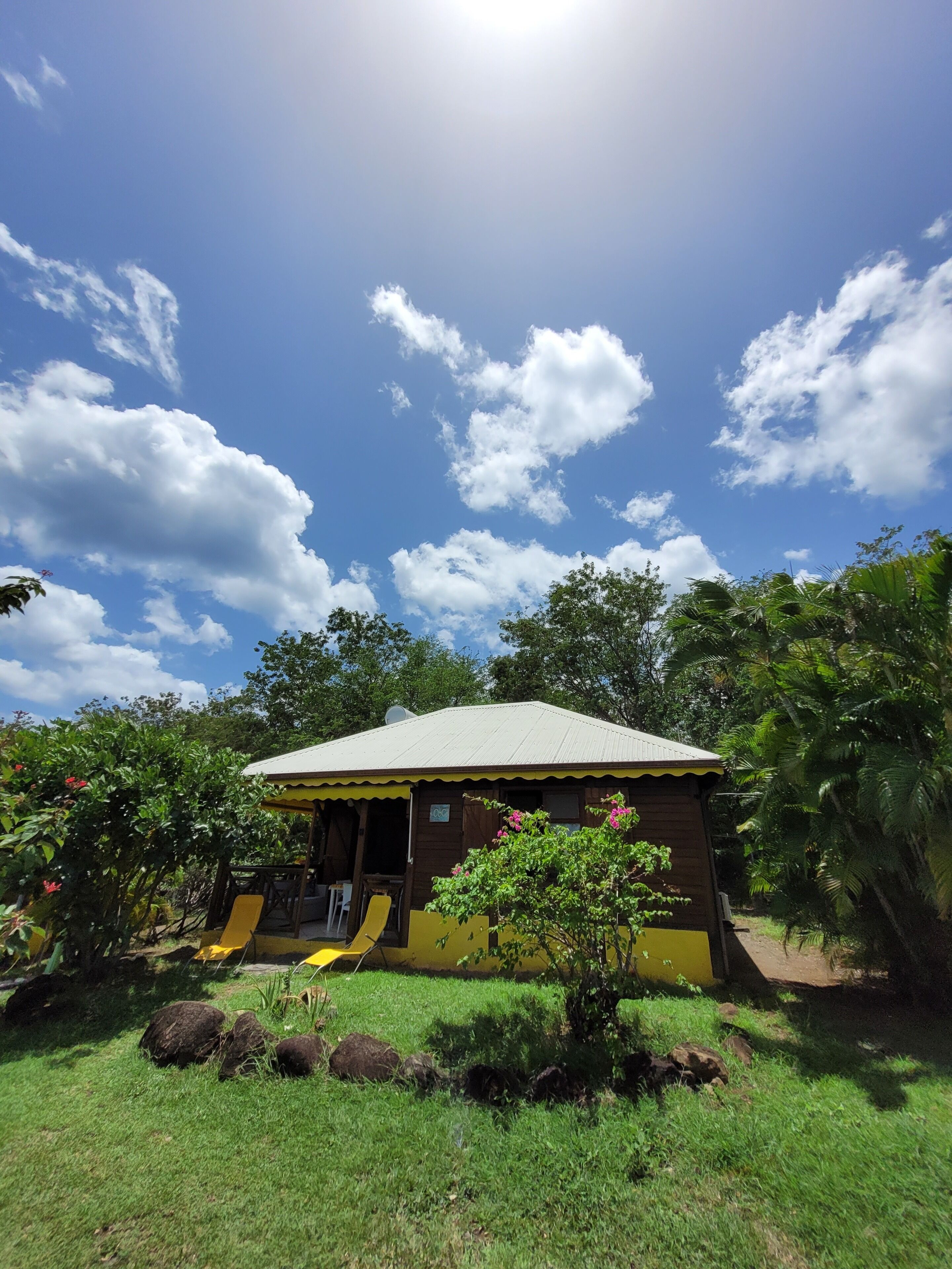 A small, single-story building with a sloped roof, surrounded by lush greenery and a clear blue sky.