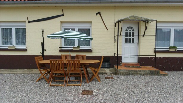 Um pátio com mesa e cadeiras de madeira, um guarda-sol listrado, e uma casa com porta e janelas brancas.