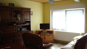 Uma sala de estar com uma mesa de jantar de madeira, televisão em um suporte, sofá e janela com cortinas.