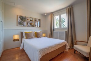 Quarto com cama, abajures, uma cadeira, janela com cortinas e obras de arte nas paredes.