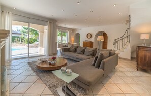 Uma sala de estar espaçosa com um sofá grande, uma mesa de centro e vista para uma piscina e jardim através de amplas portas de correr de vidro.