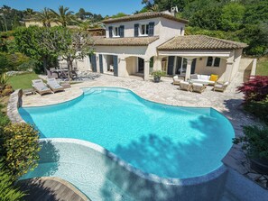 Uma piscina grande com borda curva, rodeada por um espaço de terraço com espreguiçadeiras e uma área de estar coberta.