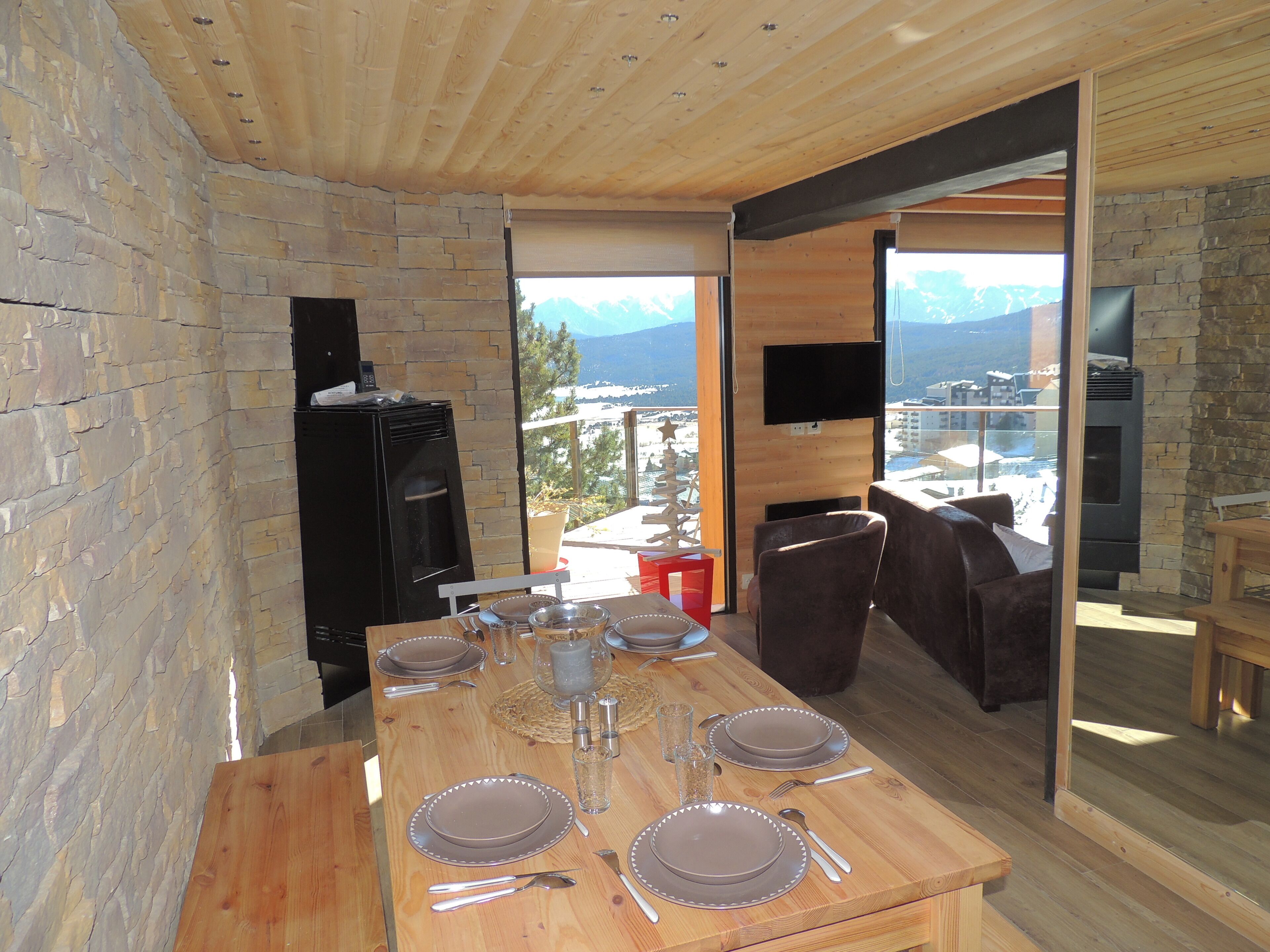 Un comedor con una mesa de madera preparada para cuatro personas, una pared de piedra, un refrigerador negro y un televisor en la pared.