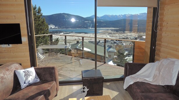 Un interno moderno con un'ampia porta scorrevole in vetro che offre una vista panoramica su un paesaggio montuoso e un lago. All'interno, due comodi divani in pelle marrone con cuscini decorativi.