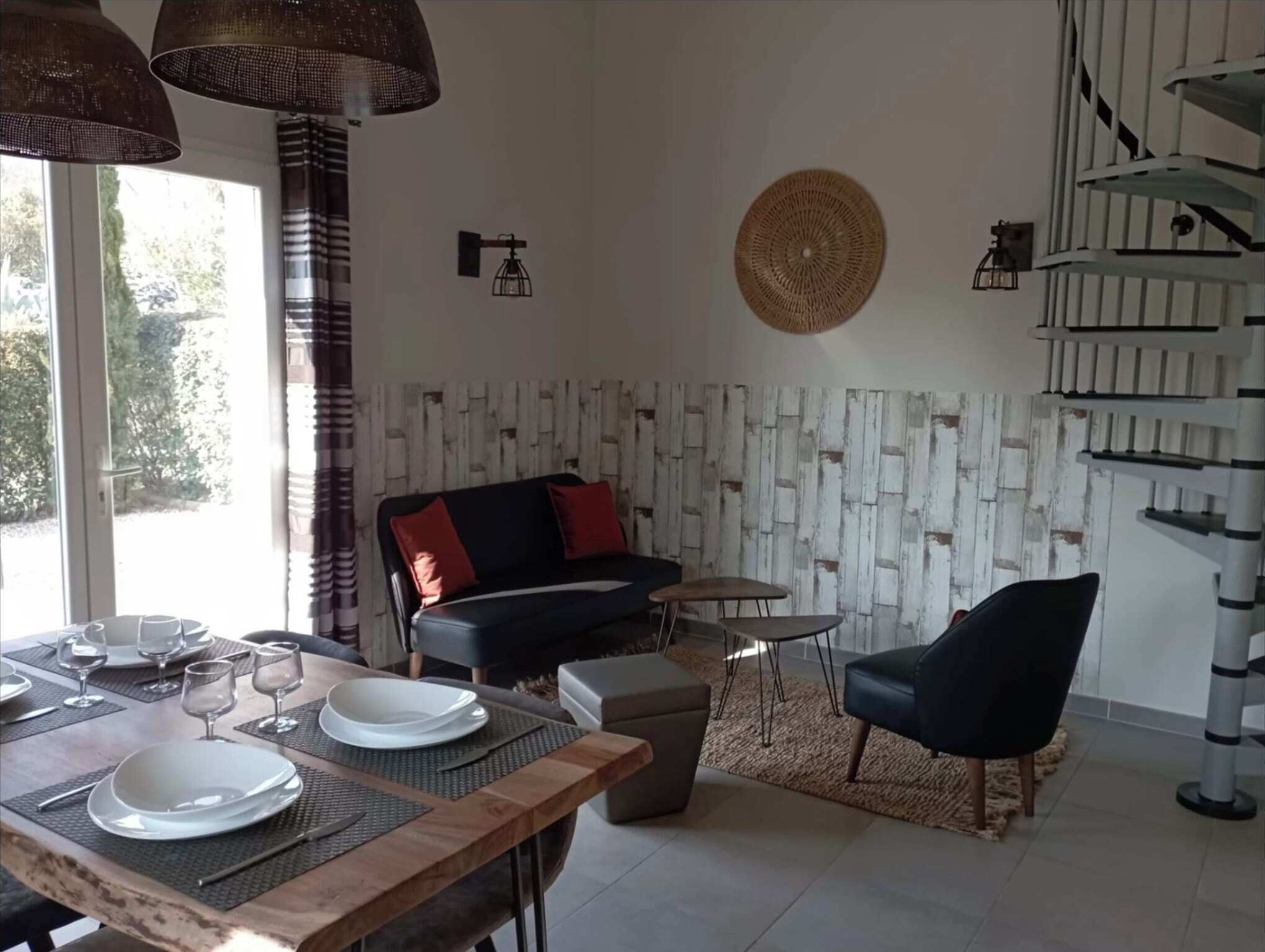 A dining area with a wooden table set for four, a black sofa with red cushions, a small round table, and a black chair.