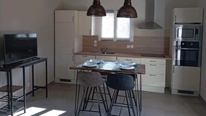 A modern kitchen with white cabinets, a wooden countertop, a dining table set for two, and a TV on a stand. - Gîte Néflier (Vogüé)