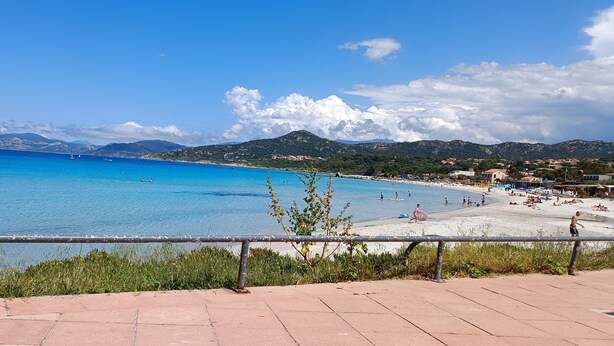 A coastal view with a clear blue sea, a sandy beach, and a mountainous backdrop.