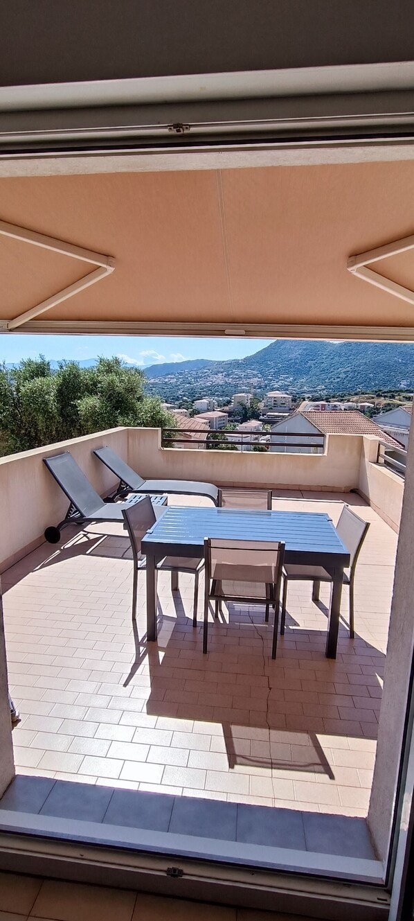 A balcony with a table and chairs, a sunshade, and a view of a town and mountains.