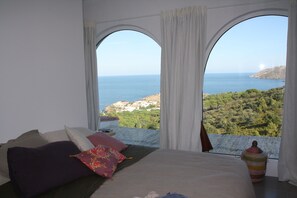Ein Schlafzimmer mit einem großen Bett, zwei Fenstern mit Vorhängen und Blick auf Meer und Grünfläche.