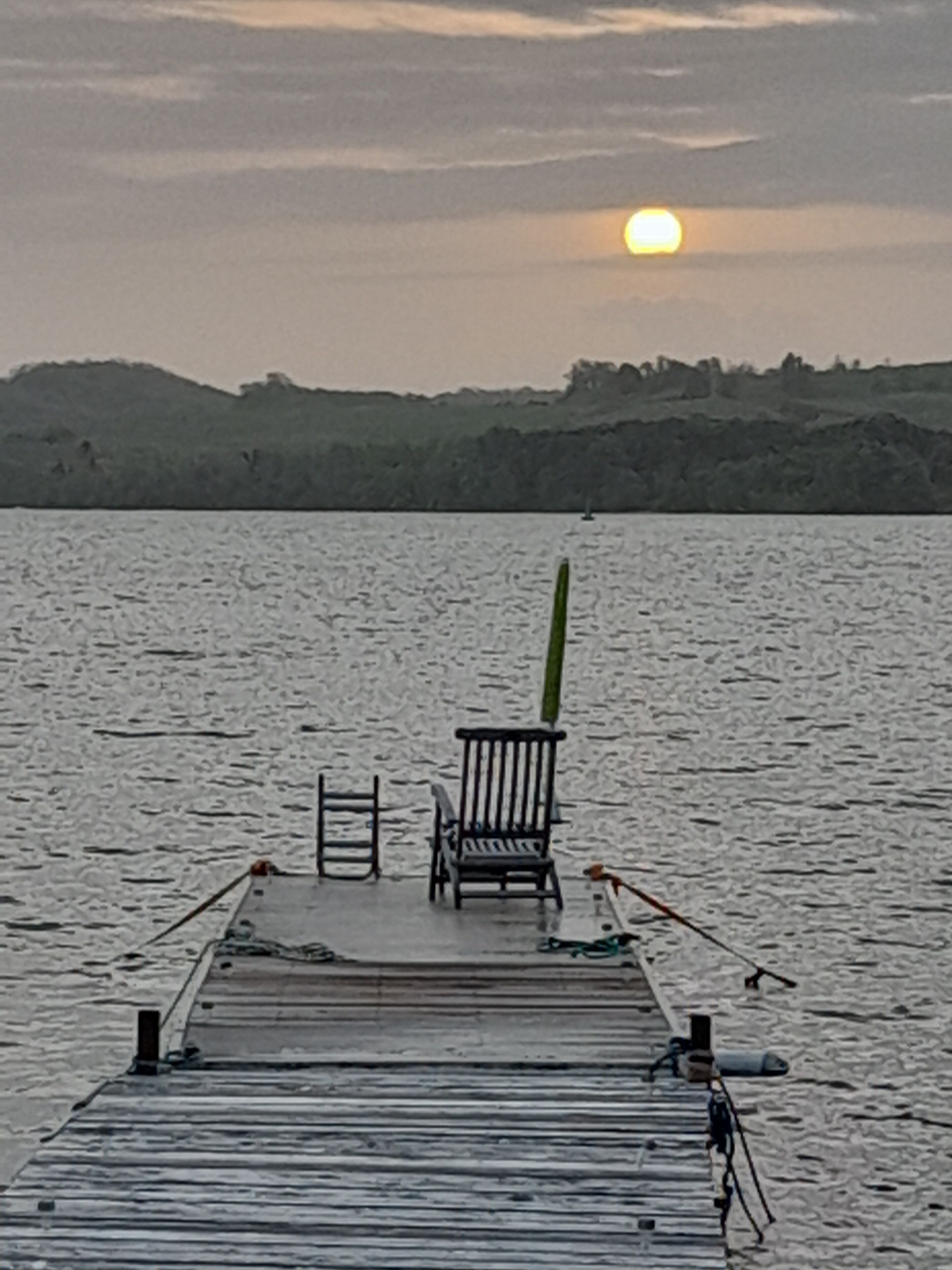 A wooden pier extending into calm water with a chair and a surfboard.
