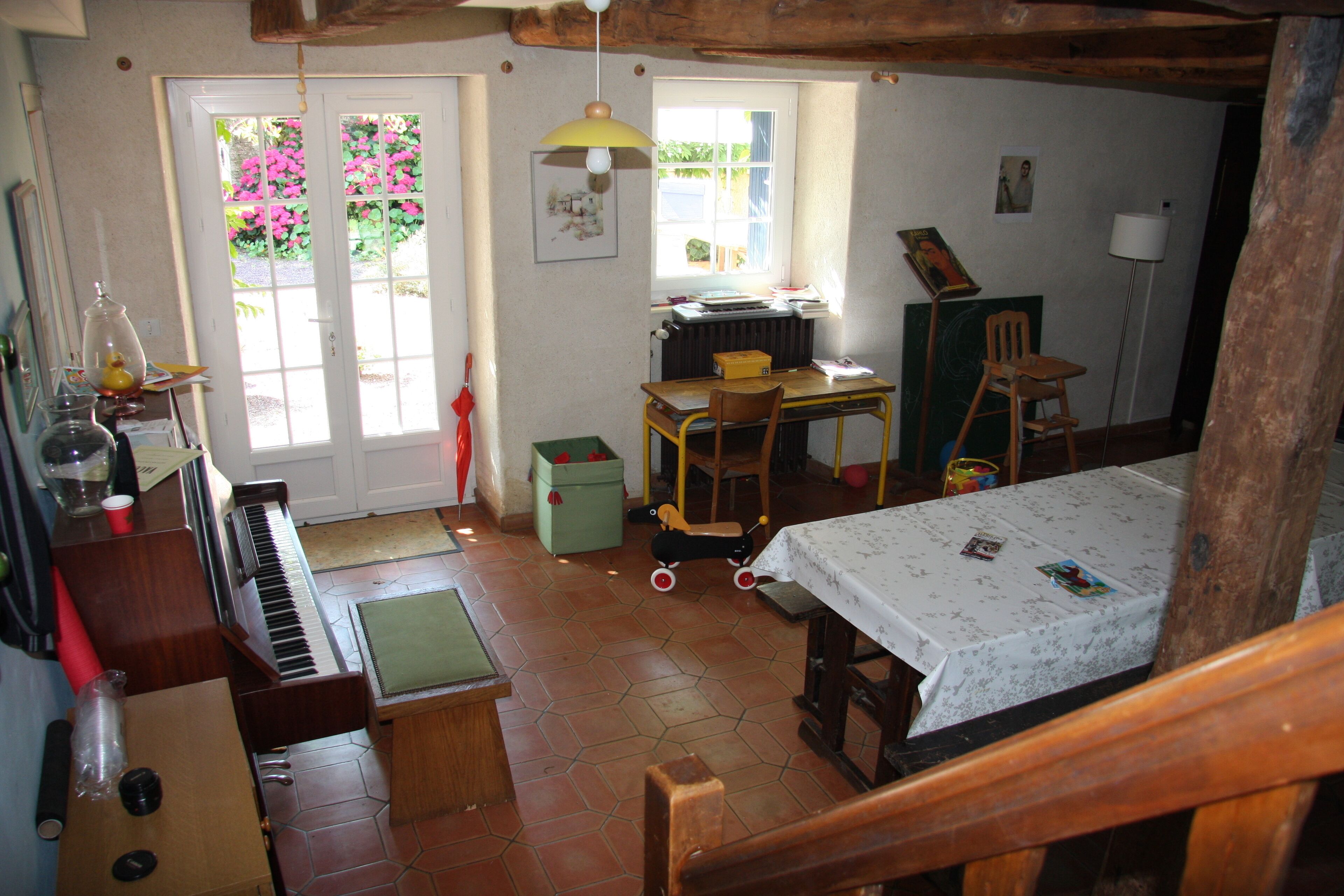 Een rustieke kamer met een piano, een tafel met een tafelkleed, een stoel, een radiator en een raam met uitzicht op roze bloemen.