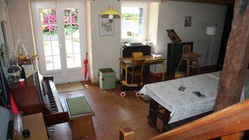 Een rustieke kamer met een piano, een tafel met een tafelkleed, een stoel, een radiator en een raam met uitzicht op roze bloemen.