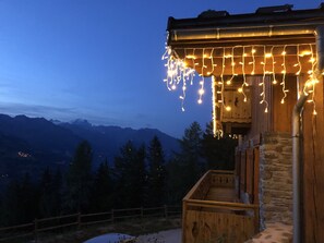 Ein Holzhaus mit einer Steinwand, einer Holzdielen-Terrasse und Blick auf Berge im Hintergrund.