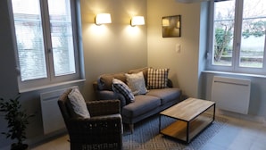 A living room with a sofa, two armchairs, a coffee table, and a patterned rug. There are two wall-mounted lamps and a framed picture on the wall.