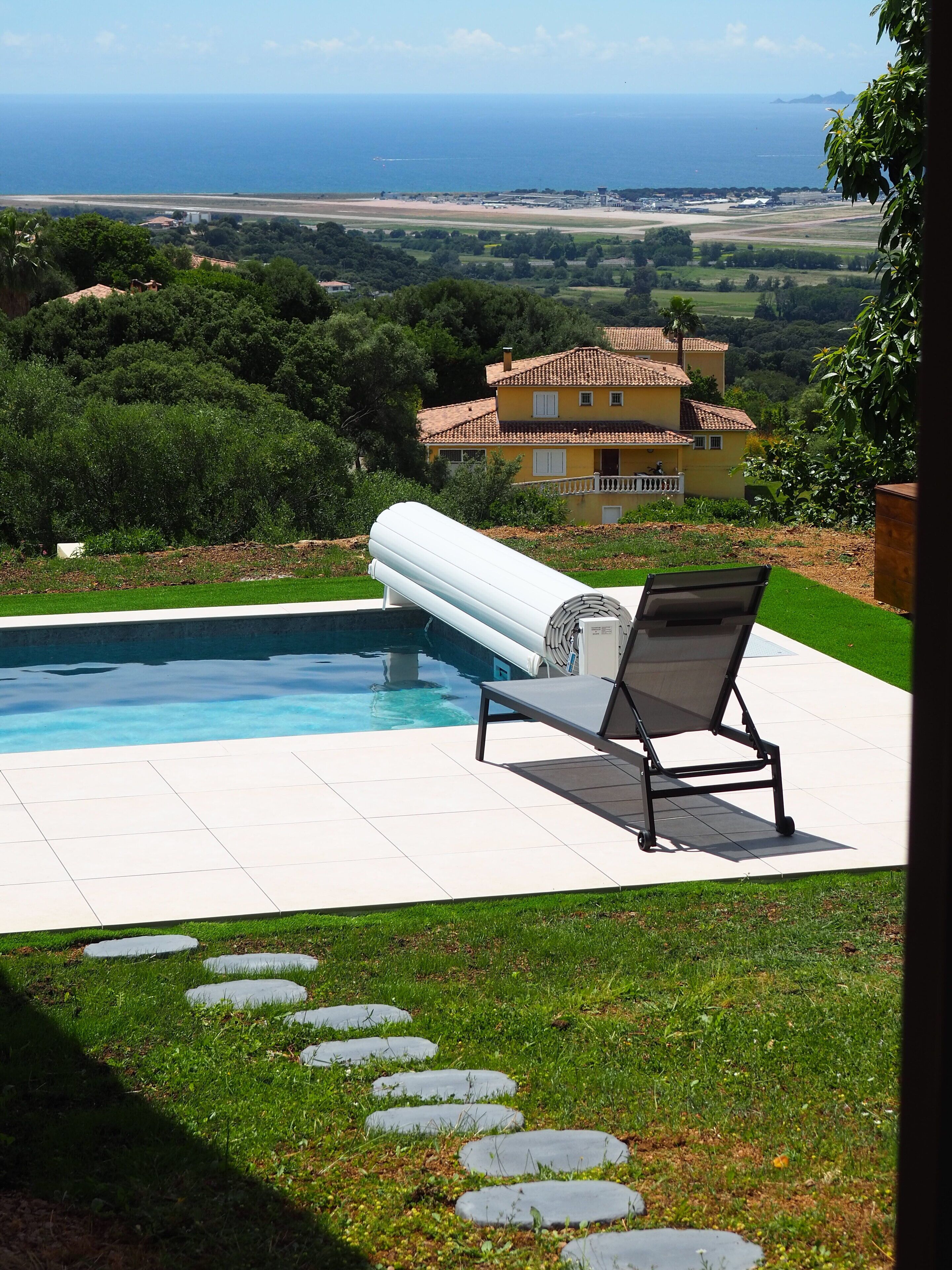 A swimming pool with a lounge chair, a stone pathway, and a view of the sea and distant buildings.