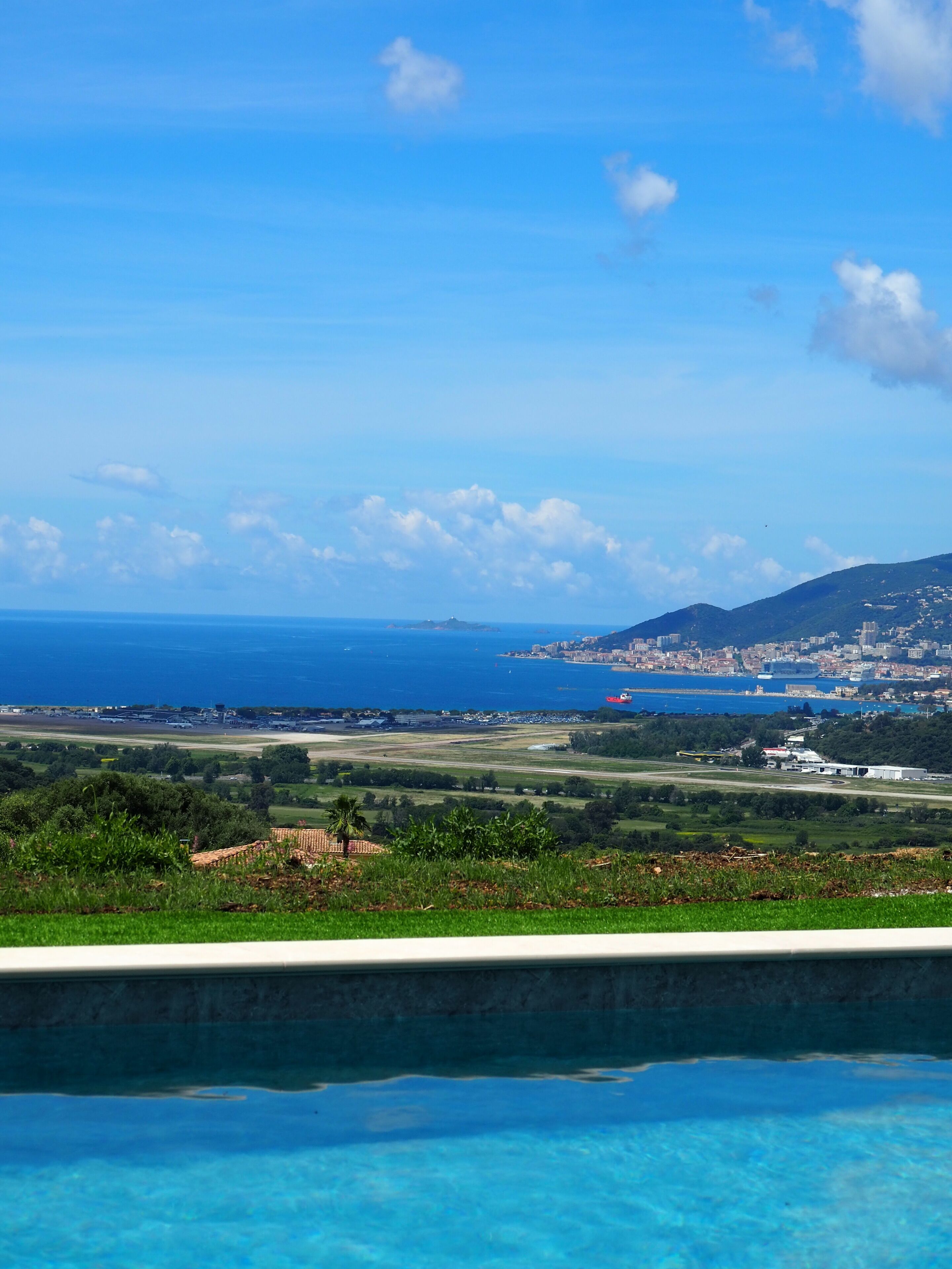 Ein Pool mit klarem, blauem Wasser, Blick auf eine Küstenstadt und eine entfernte Bergkette.