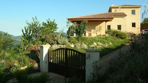 Une maison à deux étages, dotée d’un toit en tuiles, entourée d’un jardin où poussent diverses plantes et fleurs.