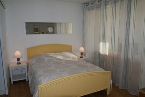 Quarto com cama, duas mesas de cabeceira, um criado-mudo, espelho e janela com cortinas.