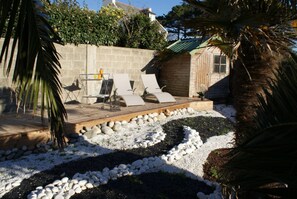Um pátio com cadeiras de descanso, uma mesa com bebidas e uma pequena área de jardim com pedriscos e pedras brancas.