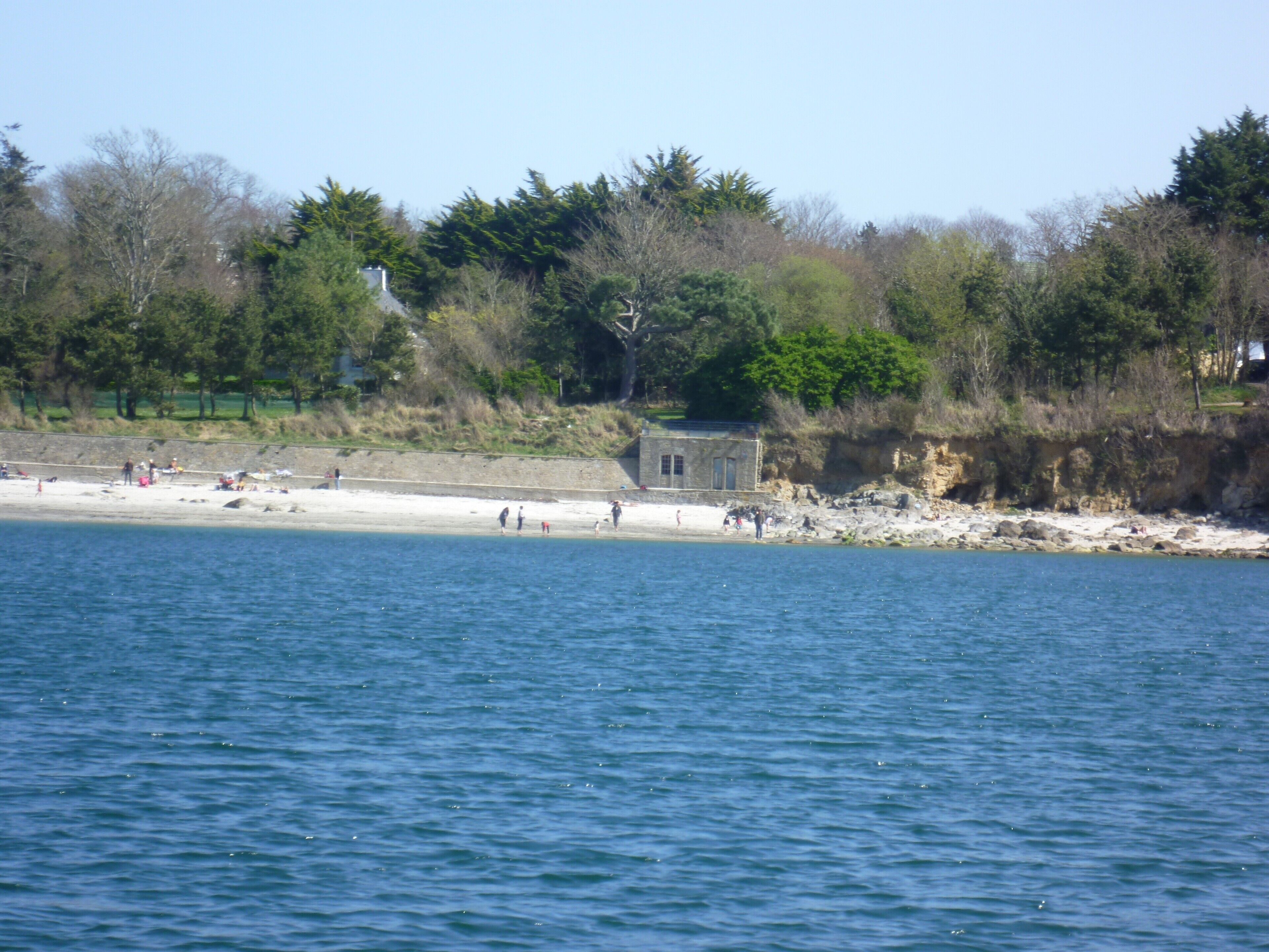 A beach with a small stone building, clear blue water, and trees in the background.