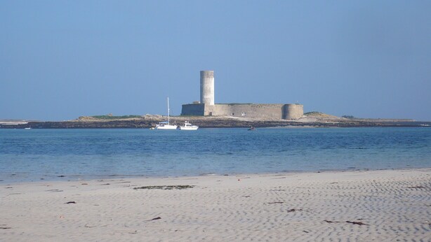 Une plage de sable avec une structure en pierre d’époque et un petit bateau sur l’eau.