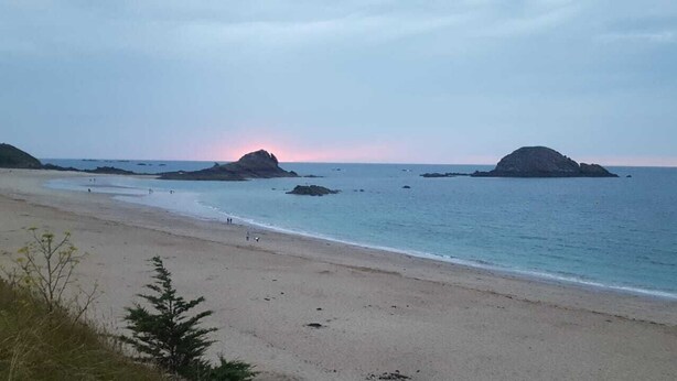 Una playa con aguas tranquilas, afloramientos rocosos a lo lejos y un cielo despejado.