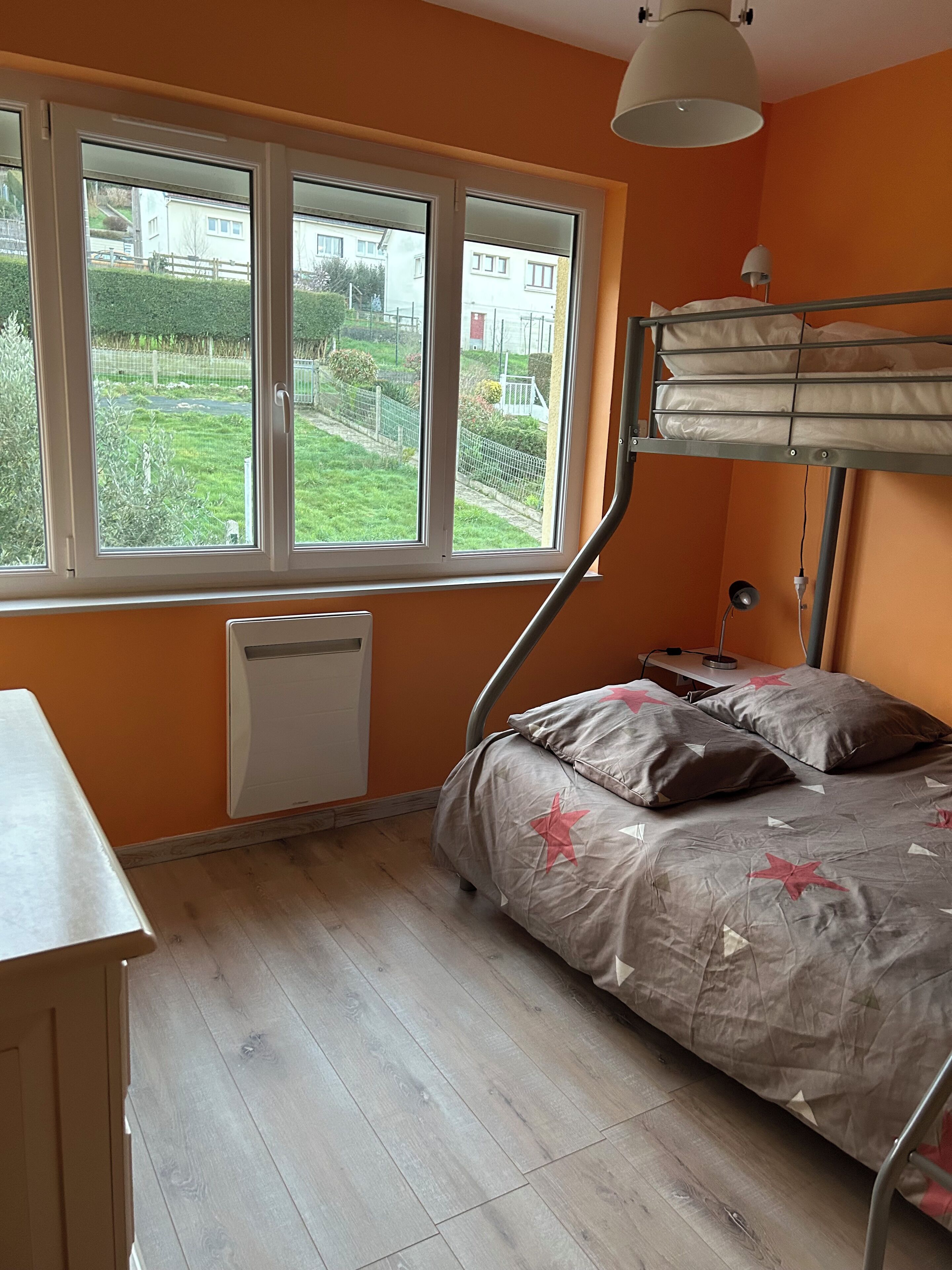 Ein Schlafzimmer mit Etagenbett, Schreibtisch, Heizkörper und einem Fenster mit Blick auf einen Garten und Gebäude.