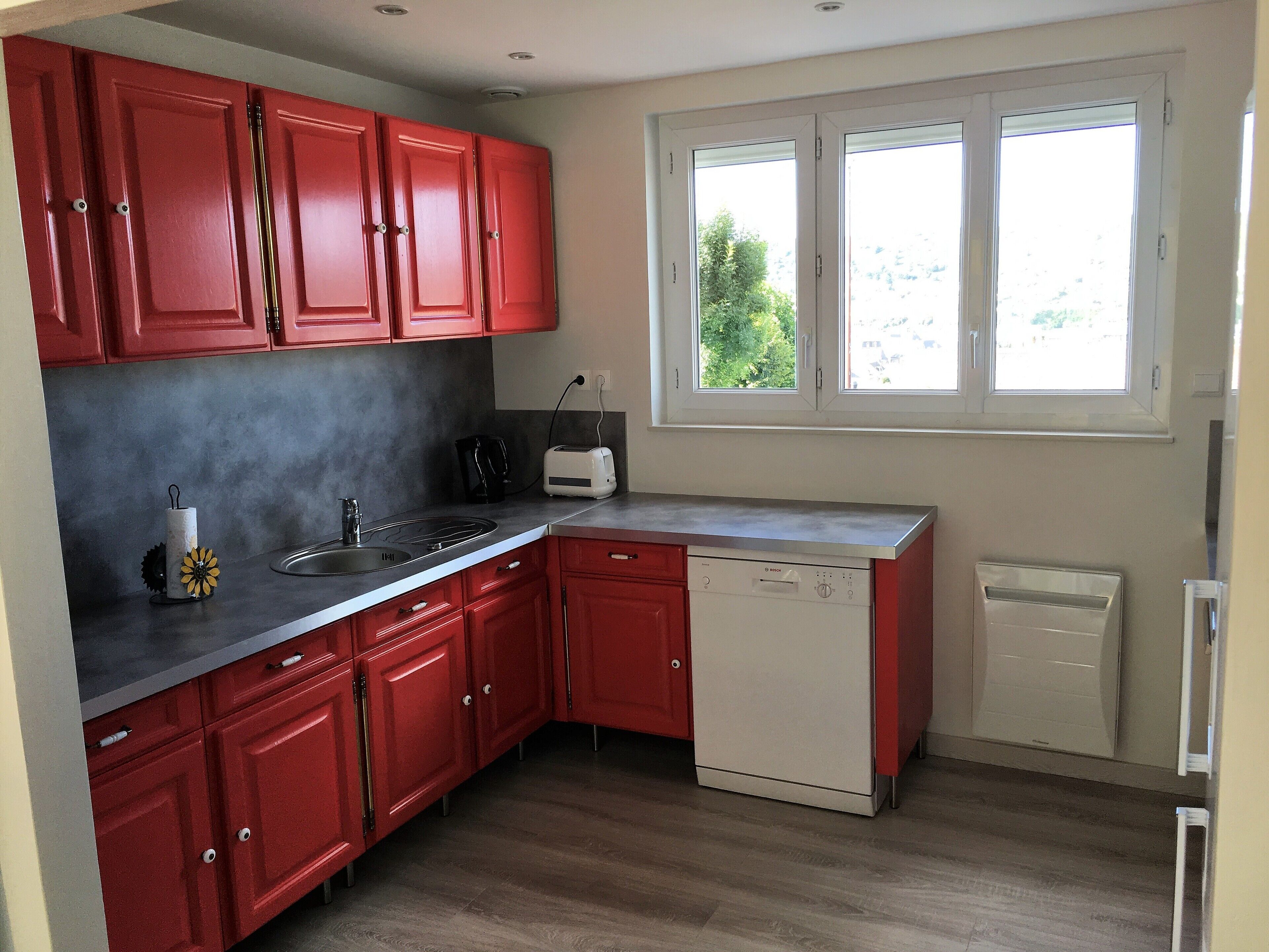 Cocina compacta con armarios rojos, lavavajillas, tostadora y ventana con vista a zonas verdes.