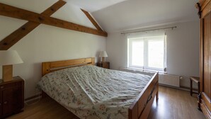 A bedroom with a wooden bed, a nightstand, a lamp, a window with curtains, and a radiator. - La Maison du Patrel - Bed and breakfast_4 people (Ban sur Meurthe/Clefcy)