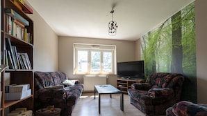 A living room with two patterned sofas, a coffee table, a bookshelf, a television, and a large forest mural on the wall. - La Maison du Patrel - Bed and breakfast_4 people (Ban sur Meurthe/Clefcy)