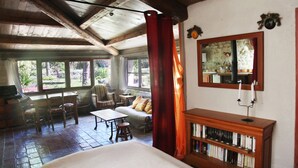 A cozy room with a large bed, a wooden bookshelf, a dining area with chairs and a table, and a view of the garden through the windows.